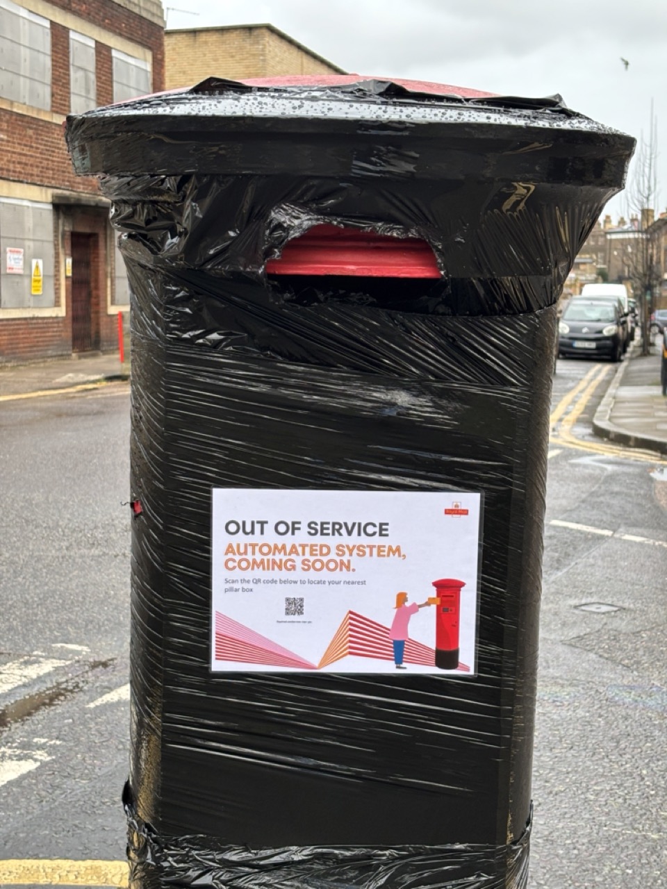 Photograph of a decommissioned post box, wrapped in black plastic, a sign reads: Out of Service, Automated System, Coming Soon